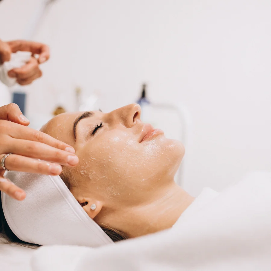 A woman receiving a facial treatment, relaxing on a spa bed with a serene expression and a therapist applying products.
