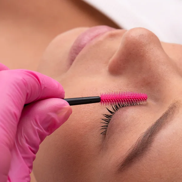Une femme se fait poser des cils avec une brosse rose, illustrant un service de maquillage permanent.