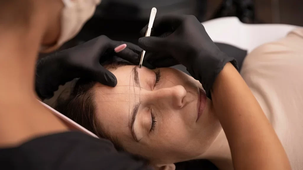 A woman receiving eyebrow micropigmentation from a professional in a beauty salon setting.