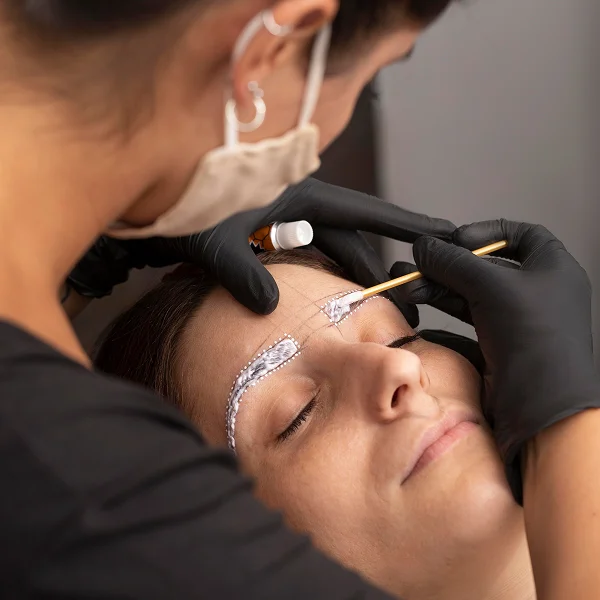 A woman receiving eyebrow micropigmentation from a professional in a beauty salon setting.